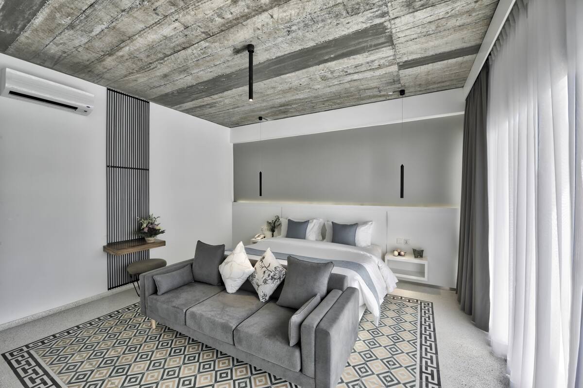 Un dormitorio moderno con un sofá gris, un tapiz estampado, una cama con ropa de cama gris y un techo con vigas a la vista.