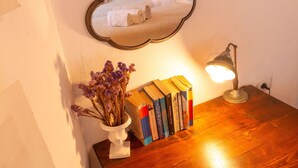 Een houten nachtkastje met een lamp, een spiegel, een vaas met gedroogde bloemen en een stapel boeken.