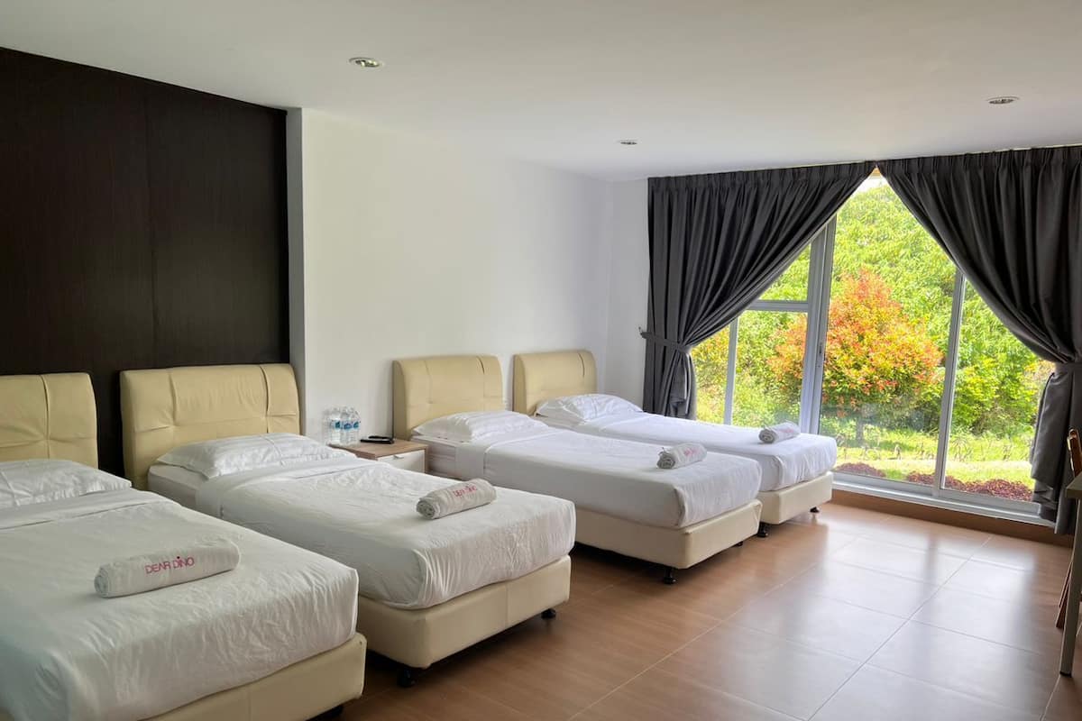 A hotel room with four beds arranged in a row, a large window with curtains, and a view of greenery outside.