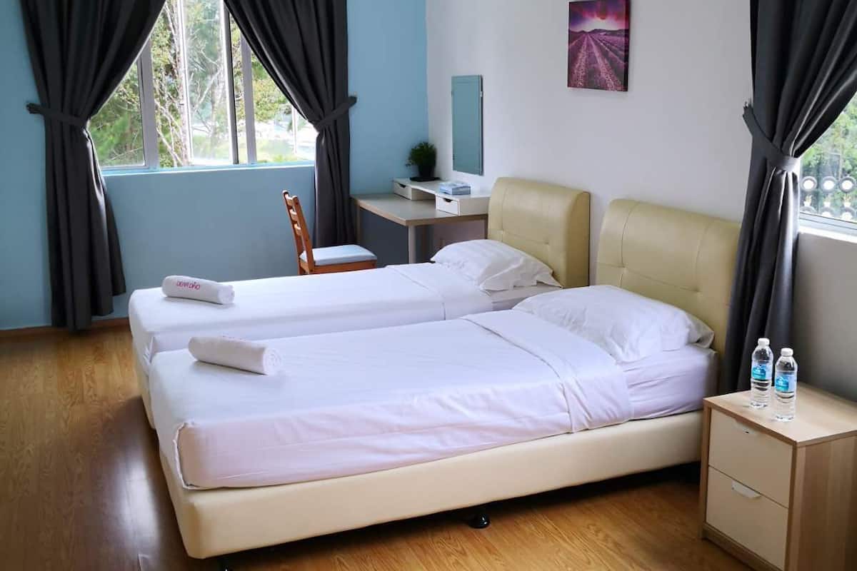 A double bed with white linens, a desk with a computer, a chair, and a small bedside table with water bottles.
