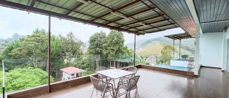 Un balcón amplio con piso de madera, una mesa de metal con sillas y barandilla de vidrio que ofrece vistas a zonas verdes y colinas.