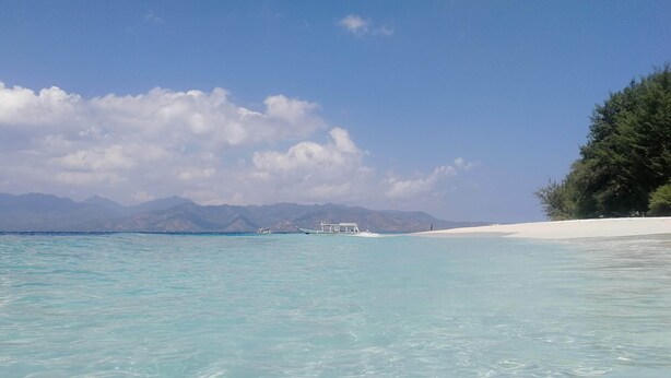 A beach with clear turquoise water, a boat, and distant mountains. - The Rabbit Tree - Hostel (Gili Meno)