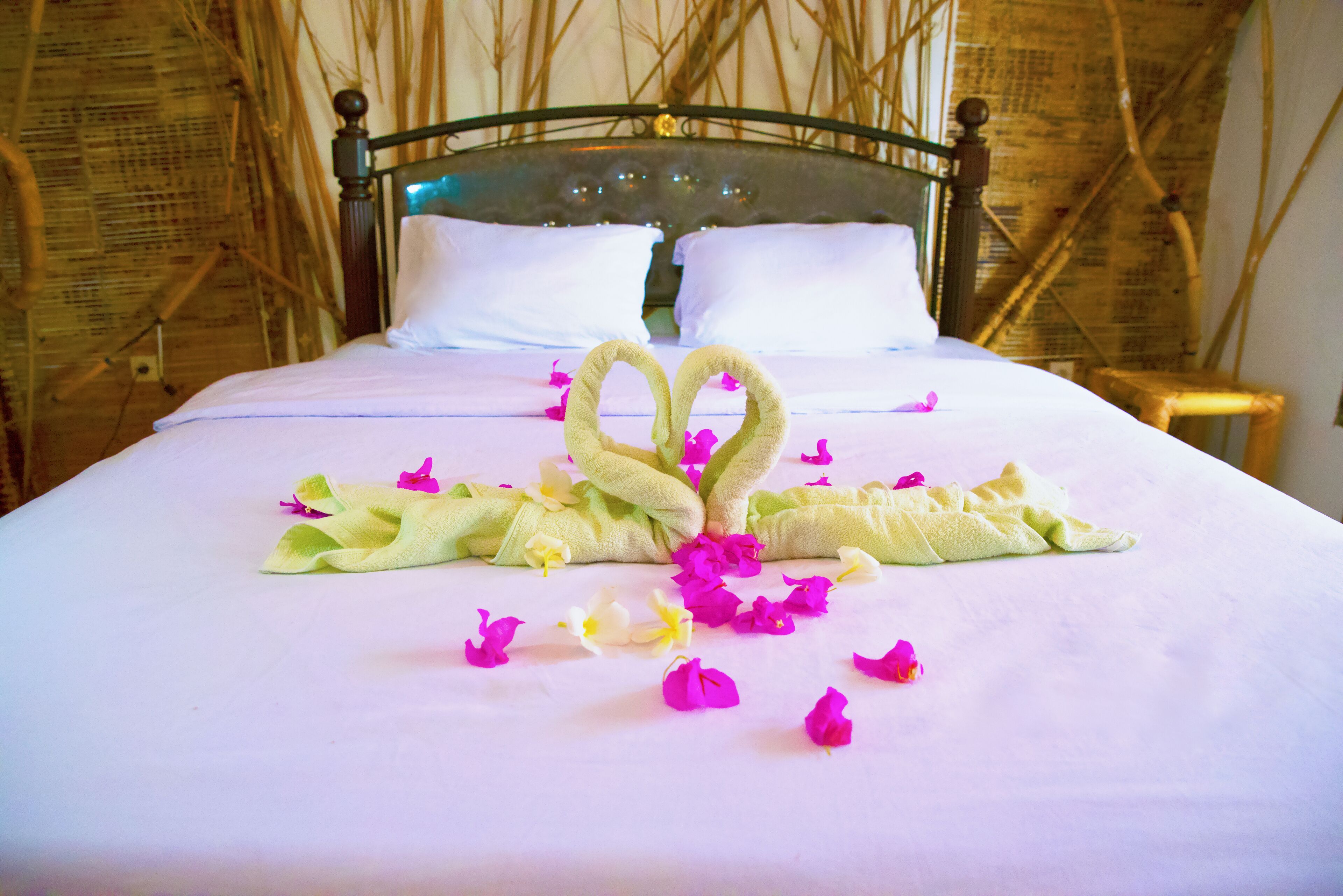 A neatly made bed with a towel arranged in the shape of a heart, surrounded by scattered flower petals.