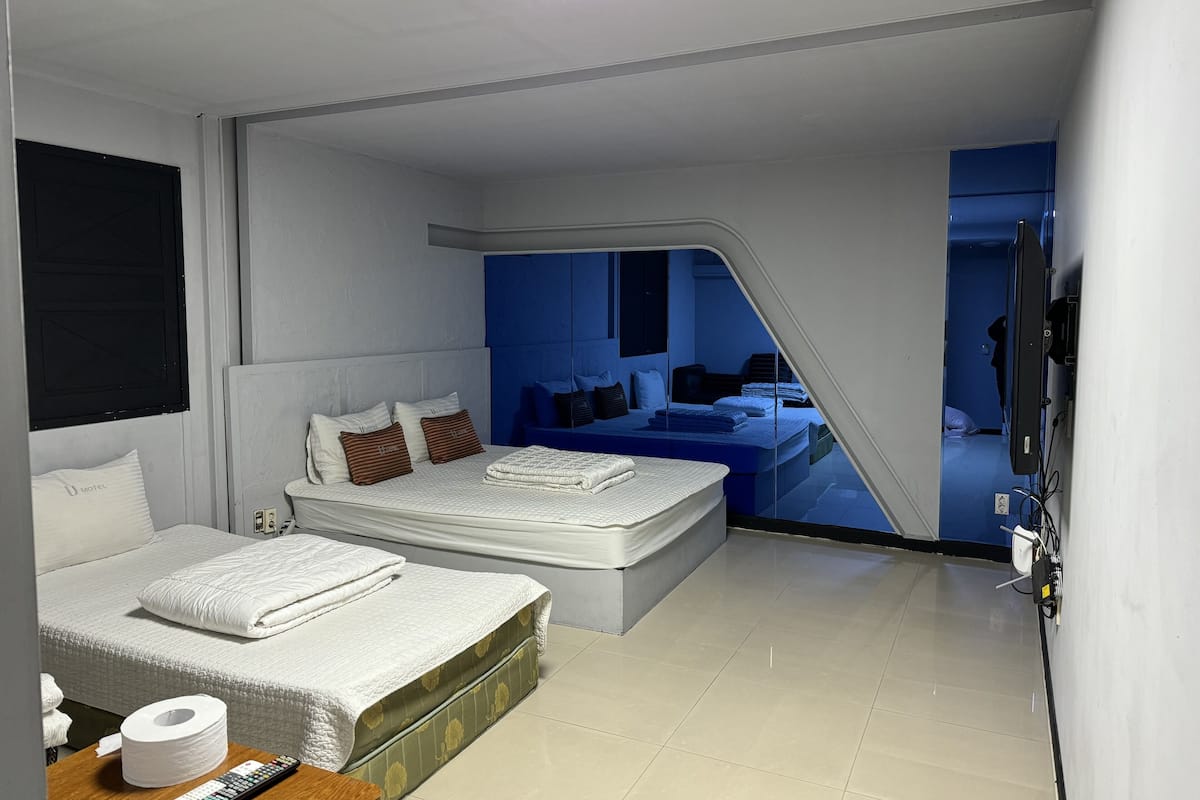 A modern hotel room with a large bed, a smaller bed, a desk with a remote control, and a bathroom visible through a glass door.