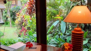 A wooden table with a book, a decorative bowl, and a lamp in a room with a view of lush greenery outside. - Alam Indah (Ubud)