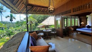 A spacious room with a large window offering a view of lush greenery. There is a seating area with a sofa and chairs, and a small table with a lamp. - Alam Indah (Ubud)