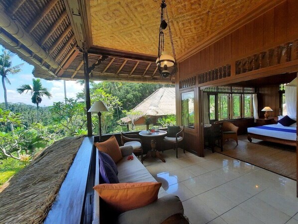 A spacious room with a large window offering a view of lush greenery. There is a seating area with a sofa and chairs, and a small table with a lamp. - Alam Indah (Ubud)