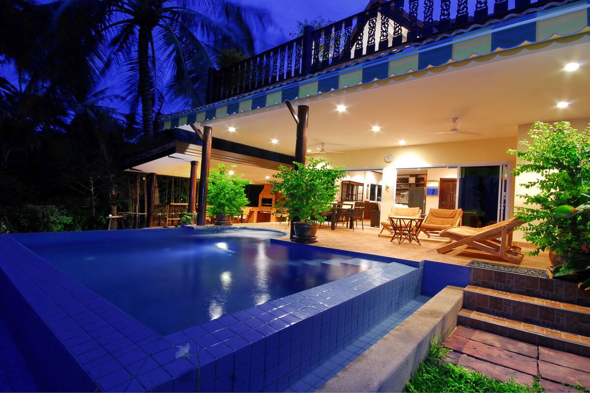 A swimming pool with a tiled edge, a covered outdoor seating area with chairs and tables, and a staircase leading to a door.