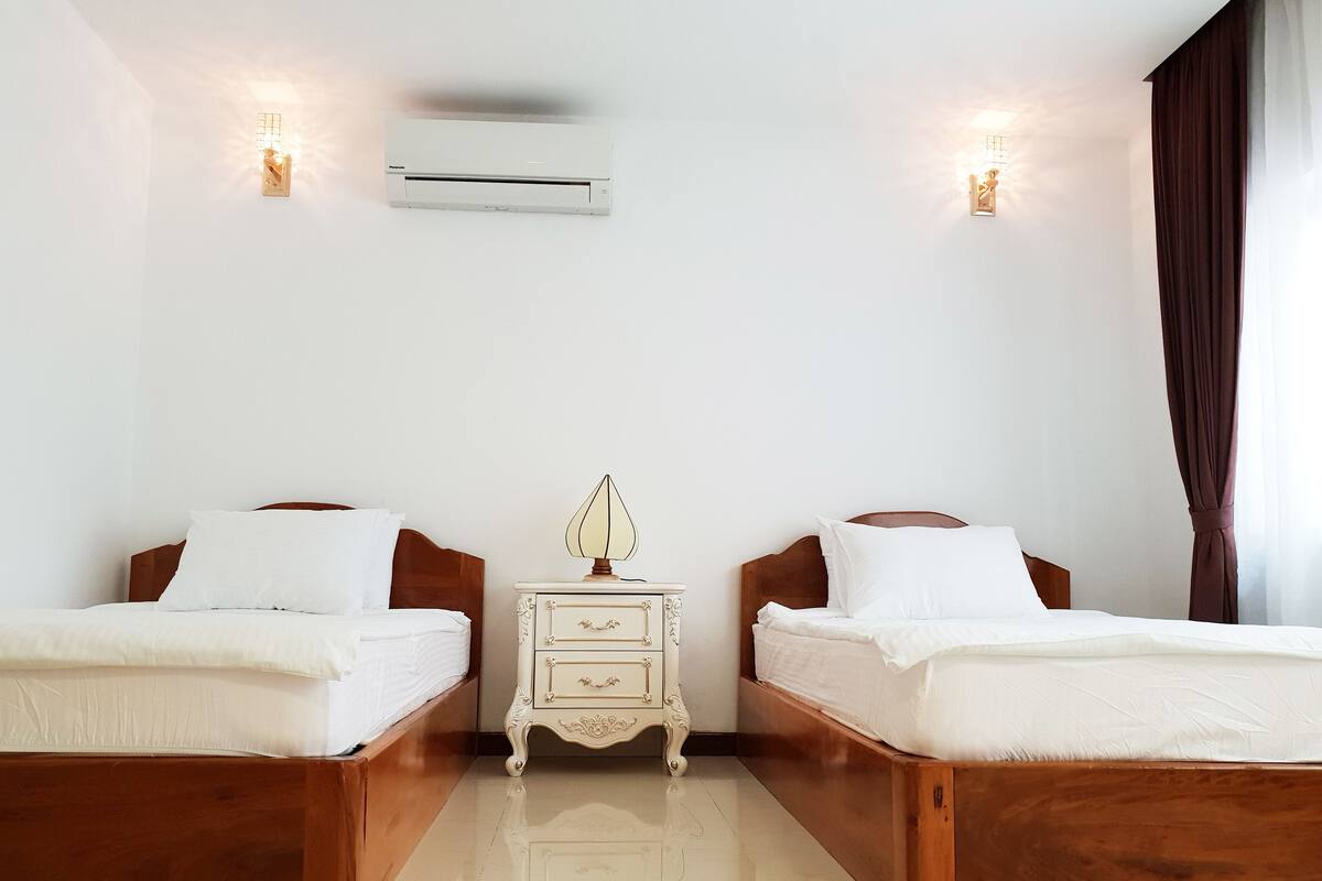 Une chambre d’hôtel avec deux lits en bois, une table de chevet avec une lampe, un climatiseur et une fenêtre avec des rideaux.