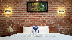 Habitació d'hotel amb paret de maó, quadre amb un bosc, dues làmpades penjades, llit ben fet amb llençols blancs i un coixí blau en forma de cor.