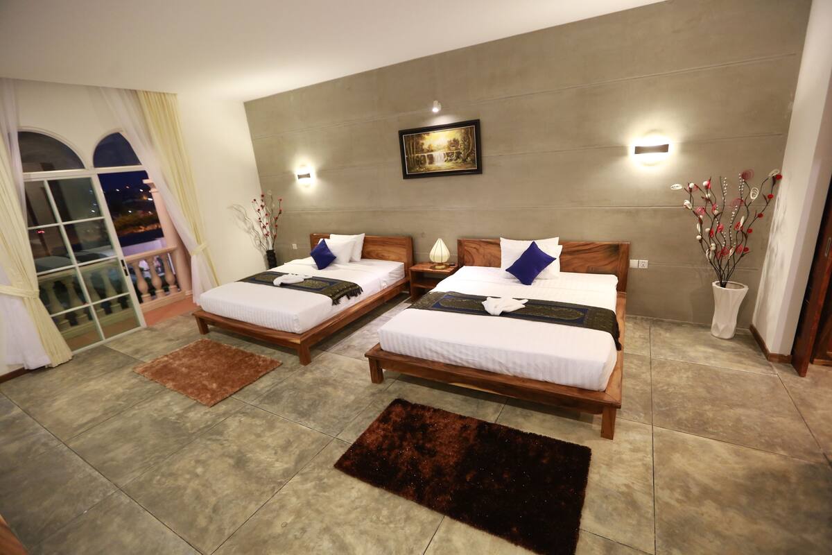 Une chambre d’hôtel avec deux lits, une grande fenêtre avec des rideaux, un tableau au mur et un vase avec des fleurs.