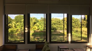 A room with large windows offering a view of a lush garden and pond. There are wooden benches and tables arranged for seating.
