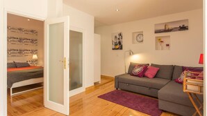 A modern living room with a grey sofa, a wooden floor, a glass door leading to another room, and wall art including a clock and a framed picture.