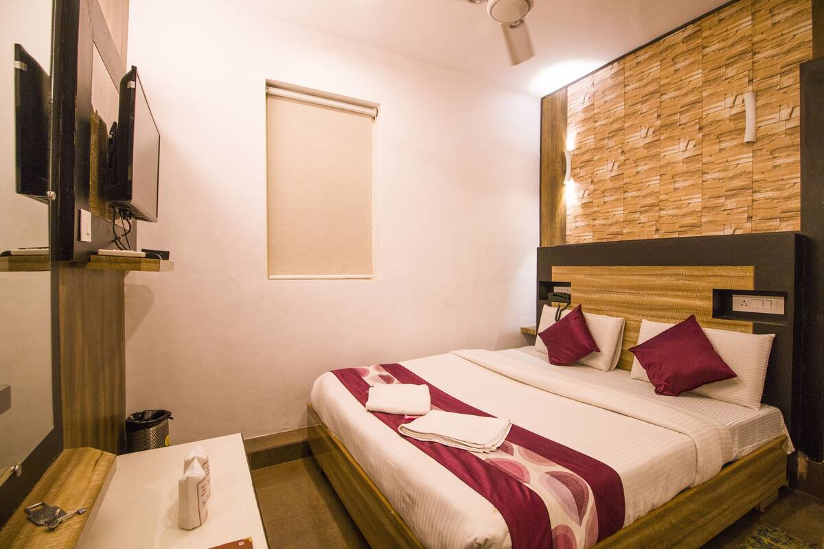 A hotel room with a wooden headboard, a bed with white and maroon bedding, a small table with a tissue box, and a wall-mounted TV.