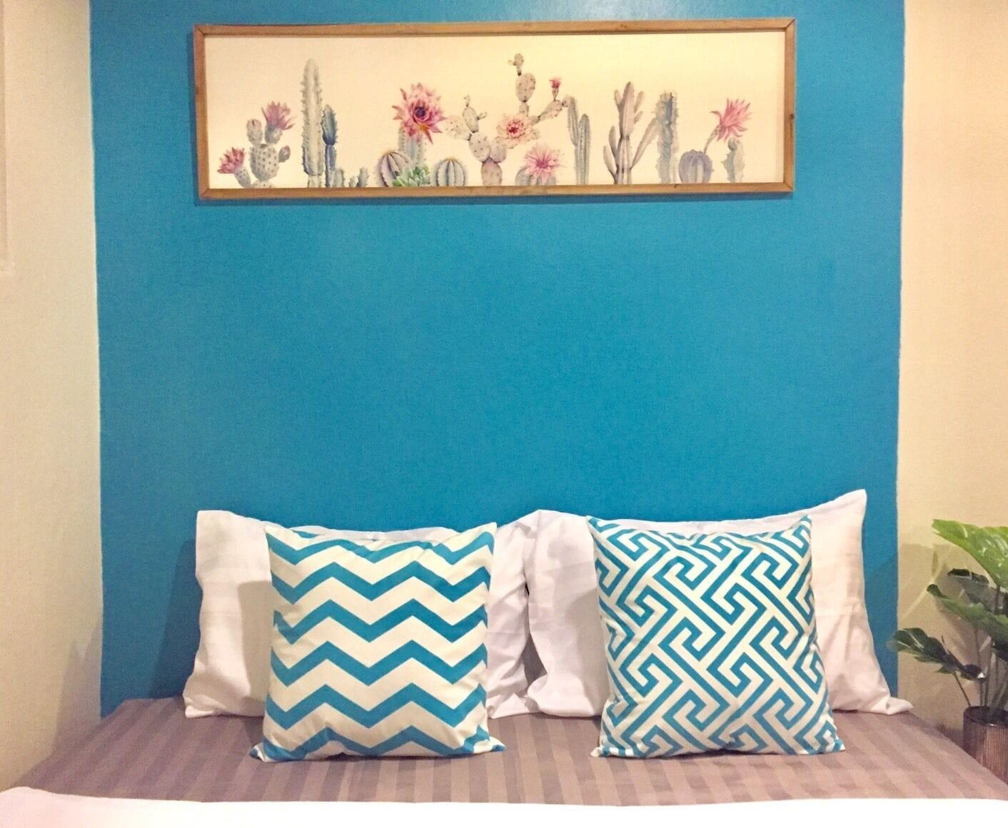 A bed with two patterned pillows, a framed floral artwork above, and a plant to the right.
