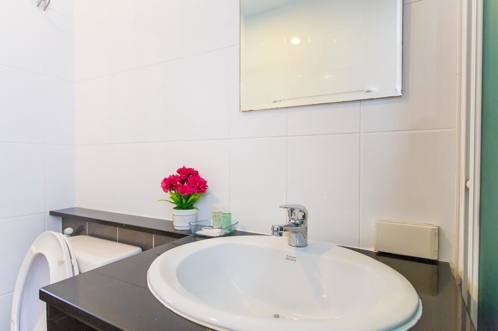 Et badeværelse med en hvid vask, et spejl, et hvidt toilet og en lille potteplante med lyserøde blomster.