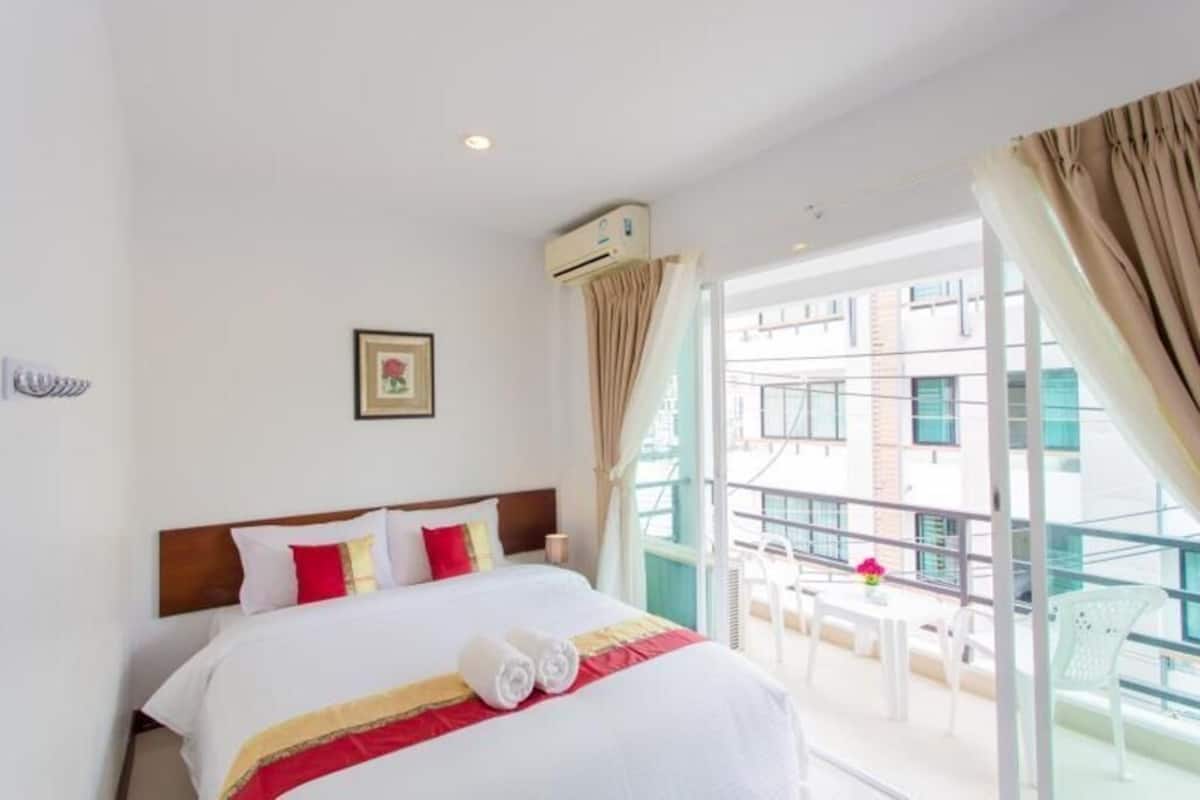 A hotel room with a bed, an air conditioning unit, a balcony with chairs, and a view of buildings.