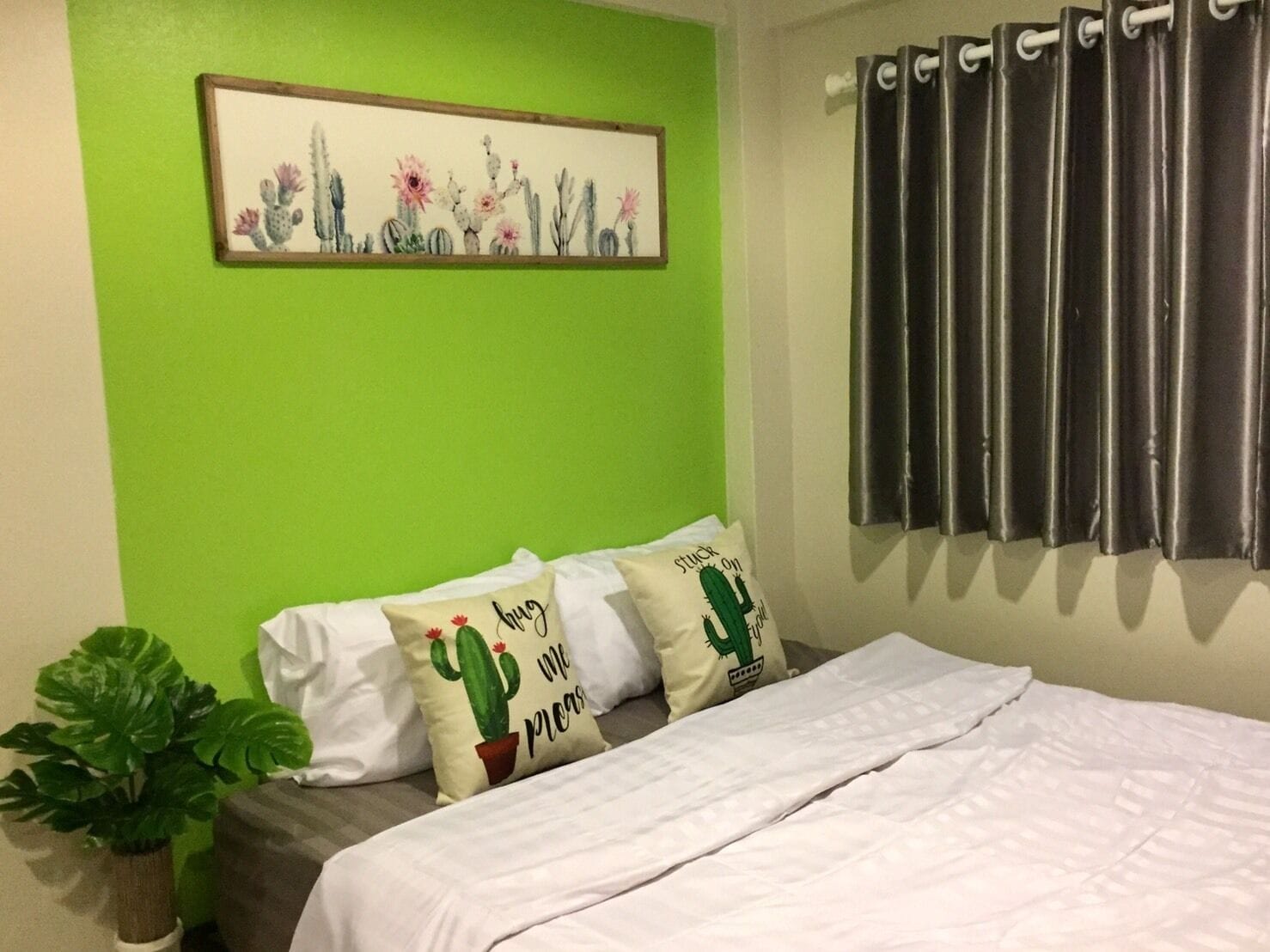 A bedroom with a green wall, a framed cactus artwork, a bed with white bedding, and two cactus-themed pillows.