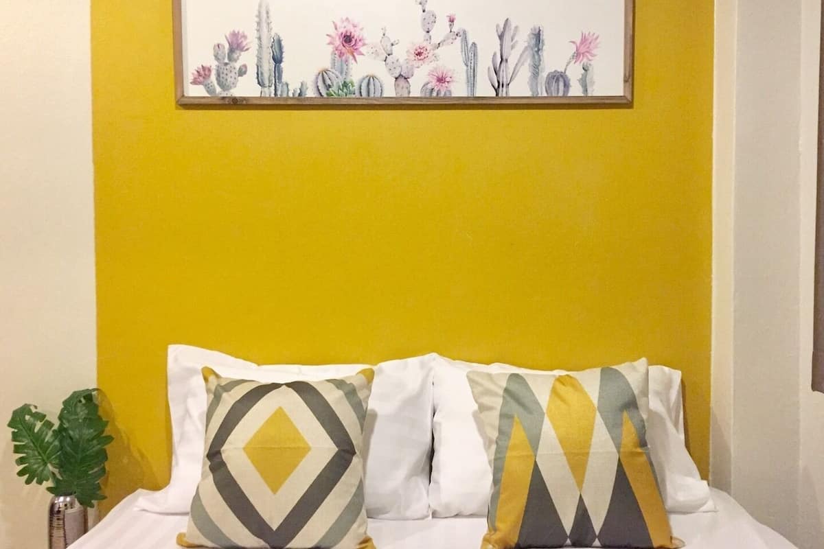 A bed with two pillows and a framed artwork of cacti and flowers on a yellow wall.