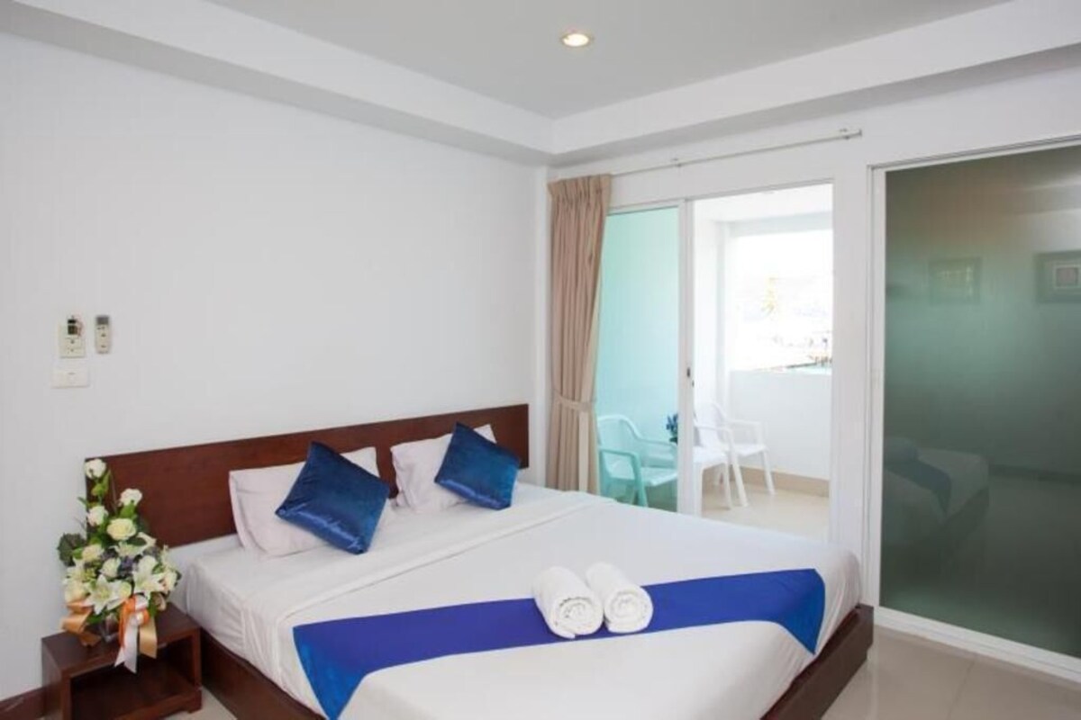 A hotel room with a bed, a nightstand with a flower arrangement, a blue bedspread, and a view of the outside.