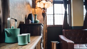 Una habitación acogedora con una mesa de madera, una tetera verde y dos tazas. Hay una lámpara con una pantalla decorativa y un sofá de cuero.