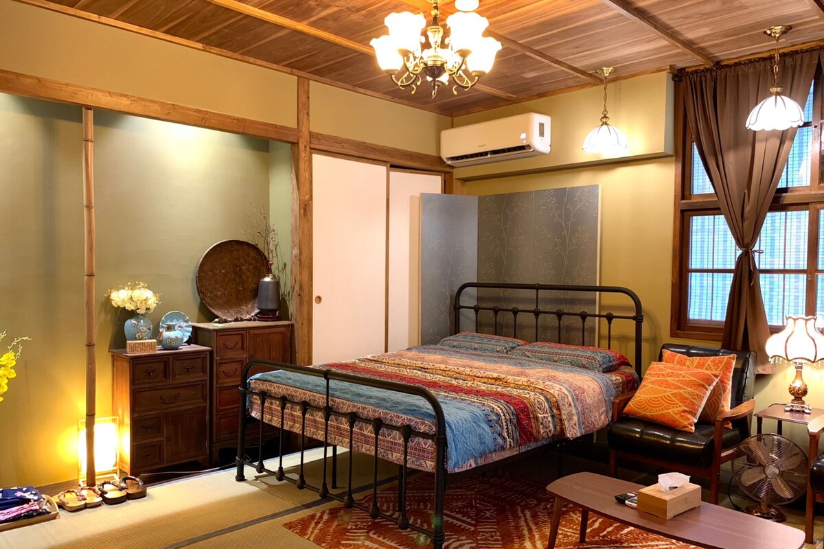 A traditional Japanese-style bedroom with tatami flooring, a low bed with a patterned bedspread, a wooden wardrobe, a small table with a fan, and a window with curtains.
