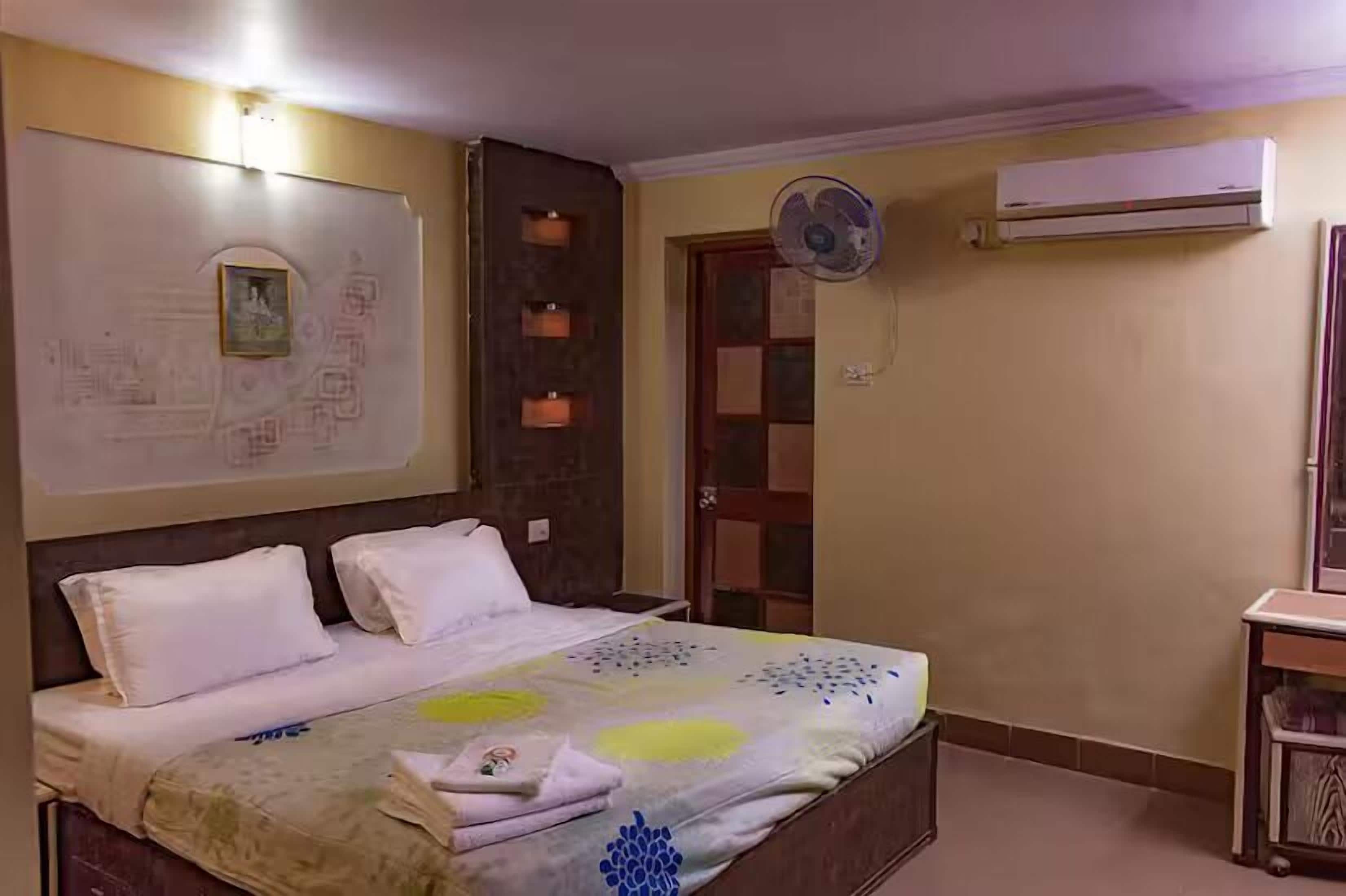 A bedroom with a bed, a wall-mounted air conditioner, a standing fan, and a door.