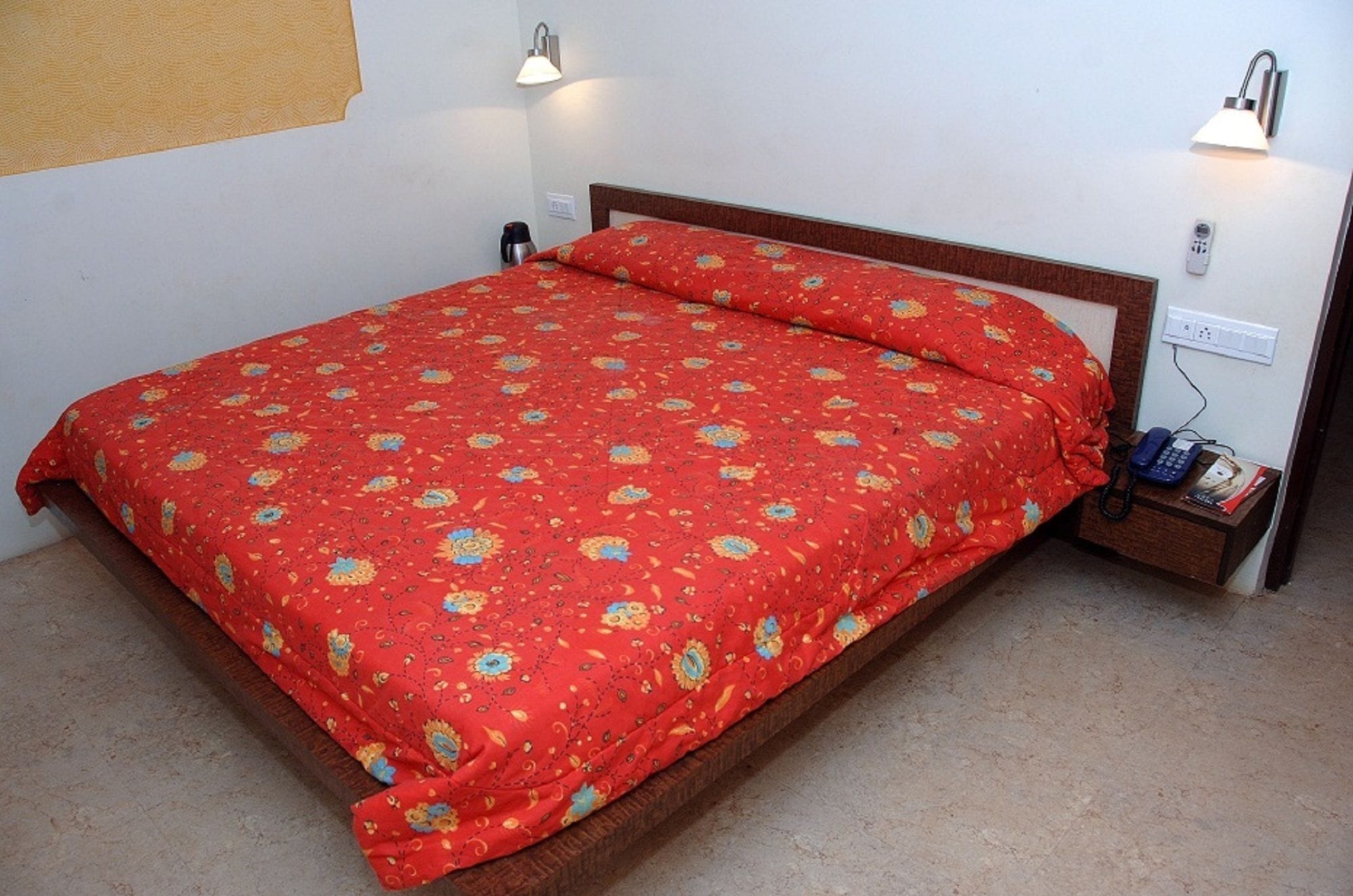 A single bed with a red floral bedspread and a wooden headboard. There is a bedside table with a lamp and a telephone.