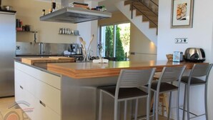 A modern kitchen with a central island, bar stools, stainless steel appliances, and a staircase leading to an upper level. - Villa Mediterranea, Urb Rio de Oro (Calonge)