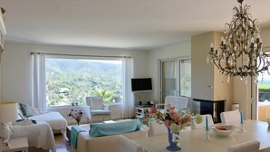 A modern living room with a large window, a chandelier, a dining table, and a TV. - Villa Mediterranea, Urb Rio de Oro (Calonge)