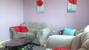 A living room with a beige sofa, a glass coffee table, and a red and blue patterned throw blanket. There are two red flower paintings on the wall. - Tulos Apartment (Ruiru)