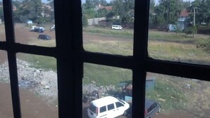 A view through a window with a grid pattern, displaying a street scene with parked cars and buildings in the background. - Tulos Apartment (Ruiru)
