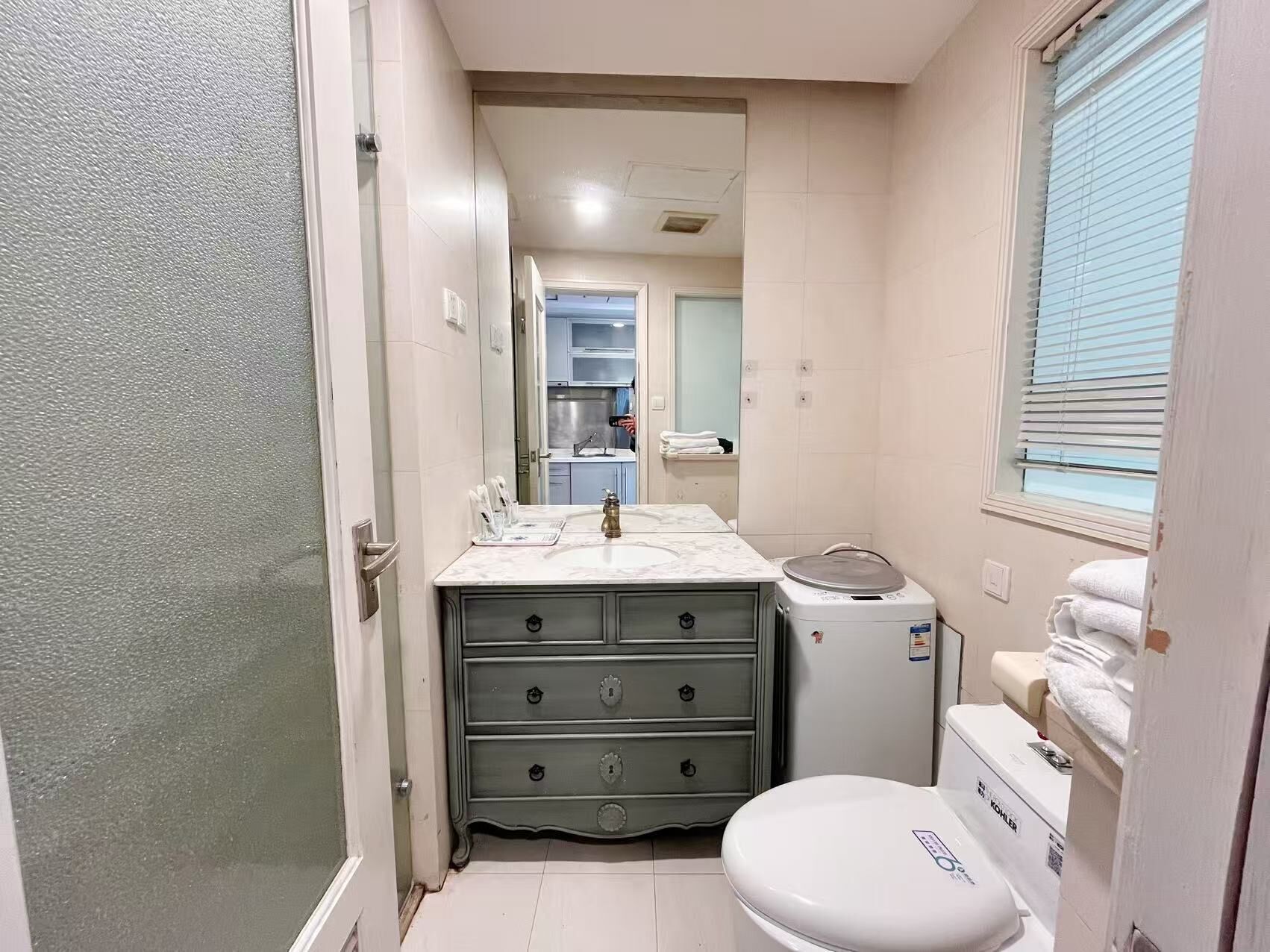 Ein kleines Badezimmer mit Toilette, einem Waschbecken mit Marmorplatte, einer kleinen Ablage und einem Fenster mit Jalousien.