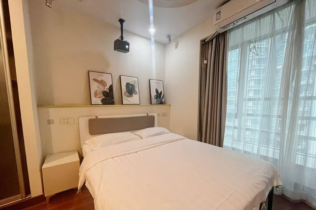 Un dormitorio con cama, mesita de noche, proyector de pared e impresiones botánicas enmarcadas.