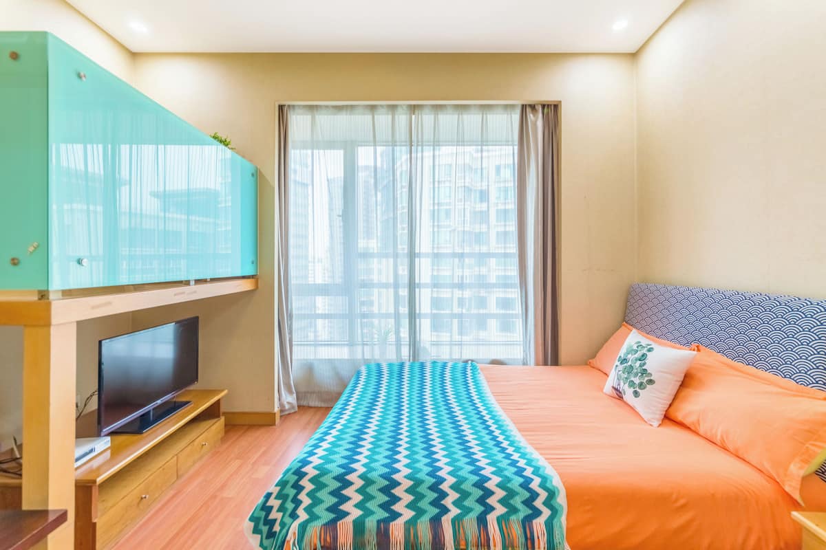 Habitación de hotel con cama, televisión sobre un mueble de madera, ventana con cortinas y vistas a edificios.