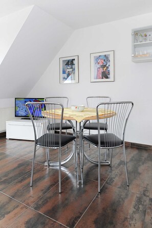 A dining area with a wooden table and metal chairs, a television on a white cabinet, and artwork on the walls.