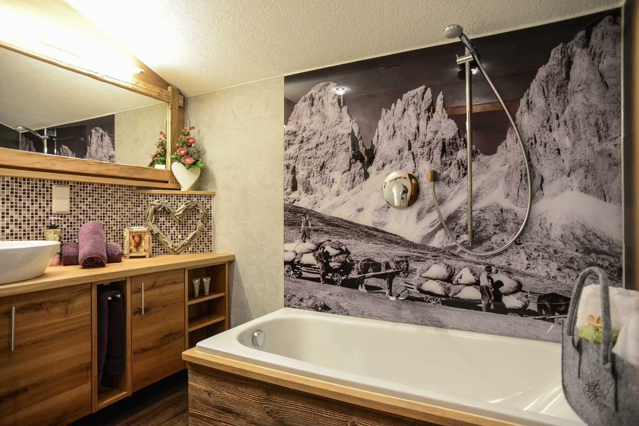 Een badkamer met een badkuip, een houten wastafelmeubel, een spiegel en een muurschildering met een berglandschap.