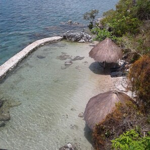 Uma piscina clara e rasa com pedras, rodeada por um muro de pedra e duas construções com telhados de palha.