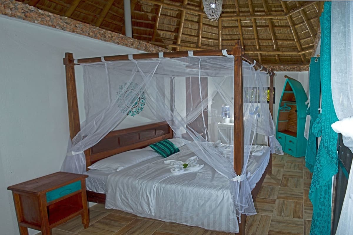 A four-poster bed with a mosquito net, a wooden nightstand, and a teal-colored wardrobe.