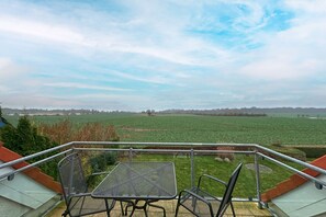 A balcony with a metal table and chairs, overlooking a green field and distant trees under a cloudy sky.