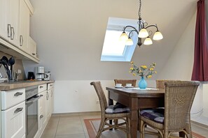 Una cocina con armarios blancos, una mesa de comedor con sillas de mimbre, un jarrón azul con flores y un tragaluz arriba.