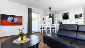 A modern living room with a black leather sofa, a round black coffee table with candles, a TV on the wall, and a kitchen area with white cabinets and a microwave. - Apartment Buhne 6 - Residence Waterkant (Esens)