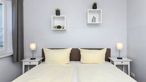 A double bed with two nightstands, each with a lamp, in a room with light grey walls. There are two white shelves with plants and decorative items. - Apartment Buhne 6 - Residence Waterkant (Esens)