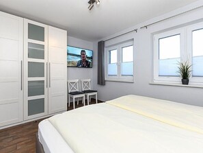 A bedroom with a bed, a TV mounted on the wall, a window with blinds, and a potted plant on the windowsill.