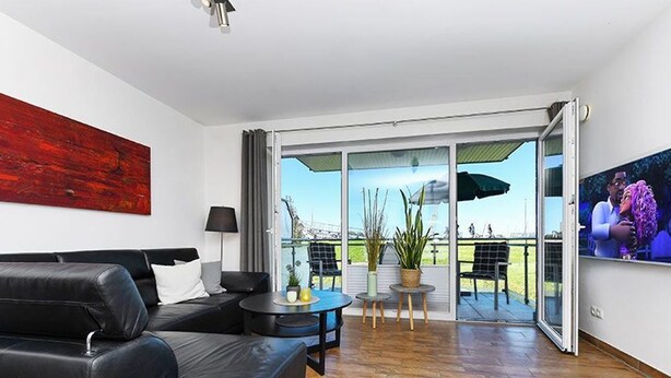 A modern living room with a black leather sofa, a small round table, and a TV on the wall. There is a sliding glass door leading to an outdoor area with a view of a park and a building. - Apartment Buhne 6 - Residence Waterkant (Esens)
