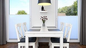 A dining area with a white table and chairs, a black pendant lamp, and a vase with flowers on a shelf. - Apartment Buhne 6 - Residence Waterkant (Esens)