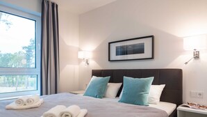 Habitación de hotel con cama, dos almohadas, mesita de noche, lámpara, cuadro en la pared y ventana con cortinas.