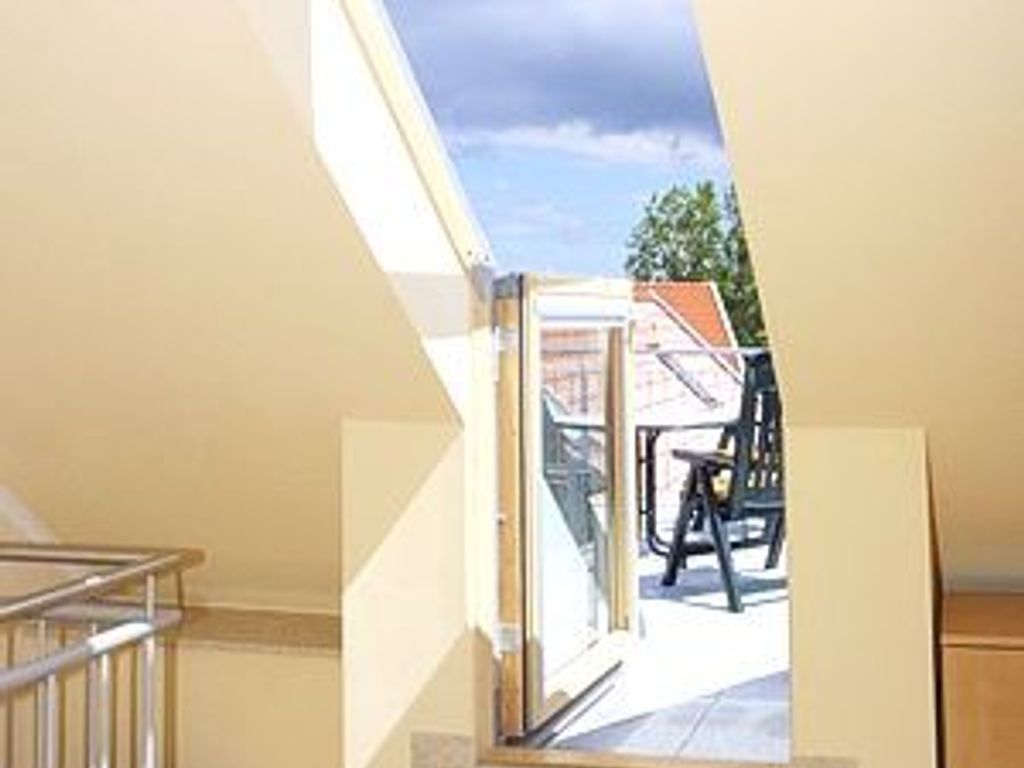 Blick aus einem Fenster auf einen Außenbereich mit Stuhl und Tisch, einem klaren Himmel und einem Gebäude im Hintergrund.