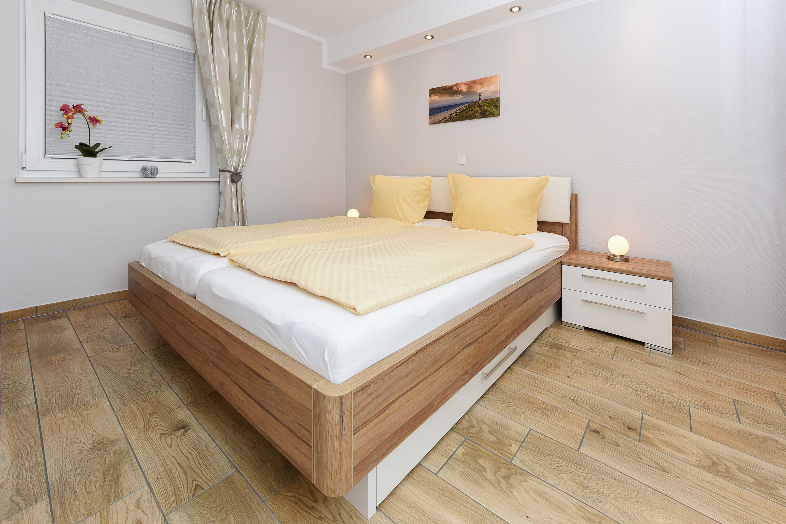 Un dormitorio con cama de madera, ropa de cama blanca, una mesita de noche con lámpara y una ventana con cortina.
