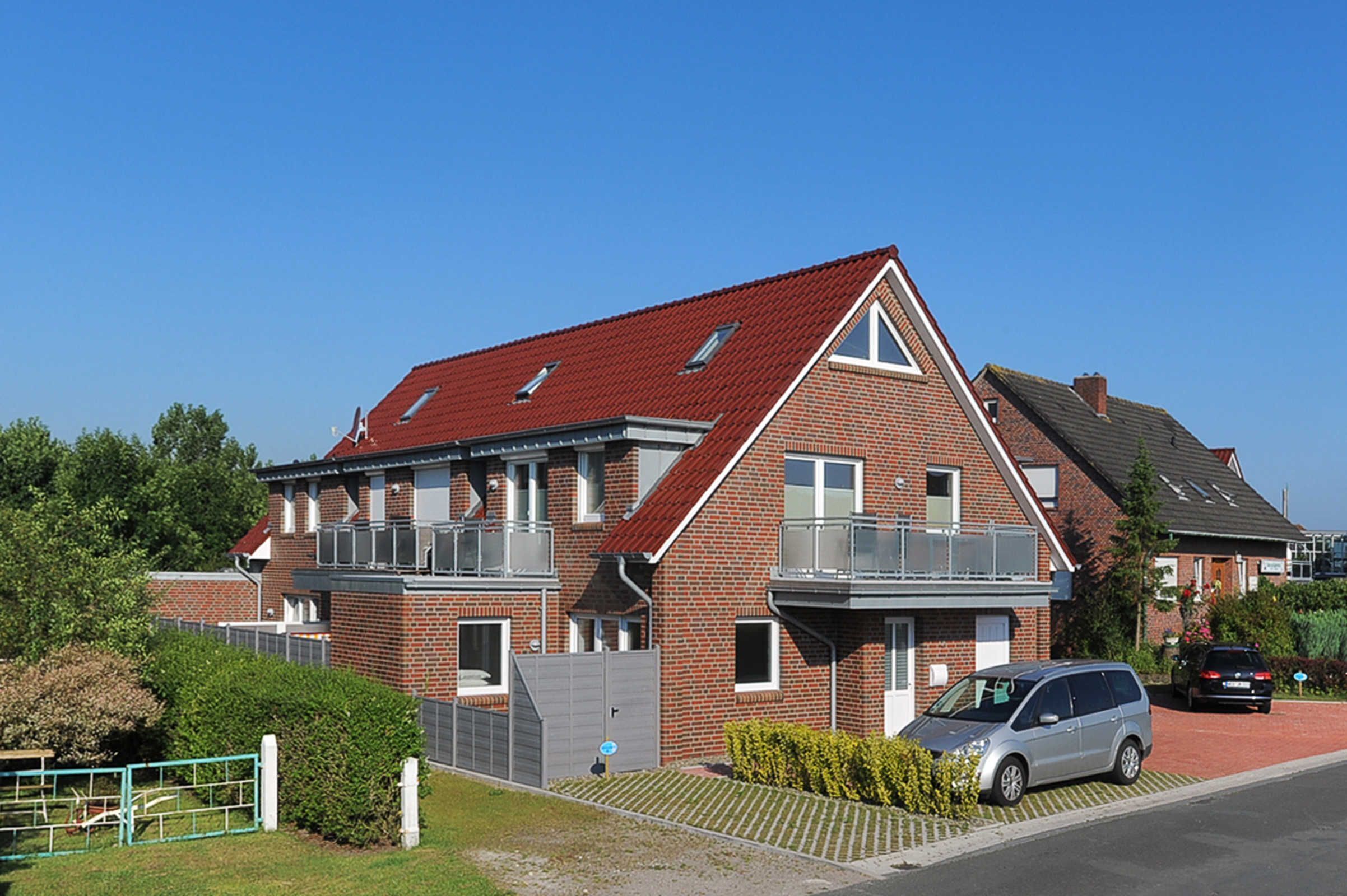Une maison à deux étages, construite en briques et coiffée d’un toit rouge, avec un balcon au deuxième étage et une voiture garée dans l’allée.